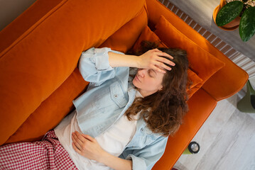Young woman lying on an orange sofa, struggling with weakness, nausea, stomach pain, and a...
