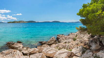 A natural rocky coastline with clear blue waters. Green trees line the shore, overlooking small islands on a sunny day.