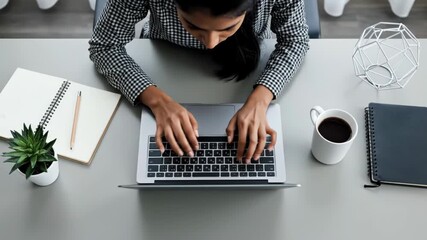 Overhead view of a focused woman typing on a laptop at a clean desk with coffee, notebook, and small plant for modern office work concept