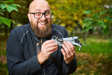 A joyful man showcases his new drone against a backdrop of lush green trees in a lively park. His excitement is evident as he holds the device with care.