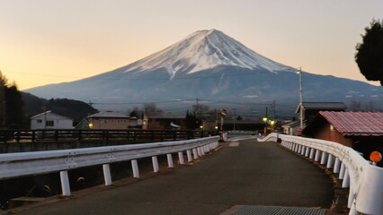 Mount Fuji rising above a quiet village near Lake Kawaguchiko at dawn, with an empty road and bridge leading toward the snow-capped mountain under a soft pastel sky