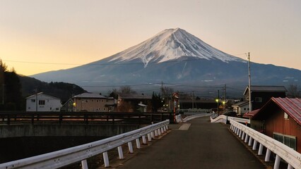 Mount Fuji rising above a quiet village near Lake Kawaguchiko at dawn, with an empty road and bridge leading toward the snow-capped mountain under a soft pastel sky