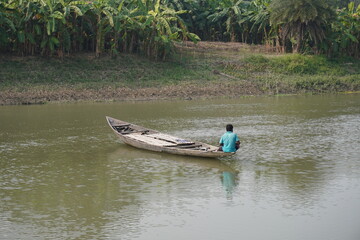 Solo fisherman in wooden boat on calm rural river with green riverside vegetation