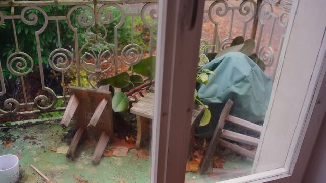 View of cluttered balcony with overturned wooden stool fallen leaves and scattered objects creating textured autumn outdoor scene seen through open window from quiet indoor perspective