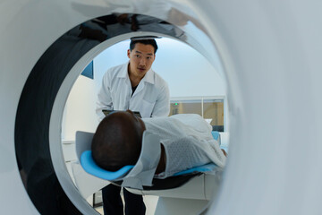 Asian doctor in coat holding tablet, African American patient lying in CT gantry with blue headrest