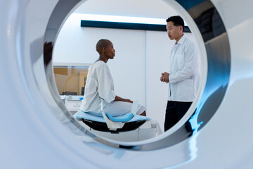African American woman patient seated in gown on gantry couch while Asian clinician explaining scan