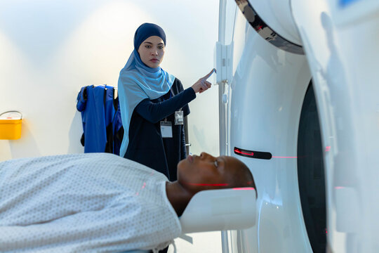 CT technologist in blue hijab adjusting CT gantry while male patient lying under red laser lines