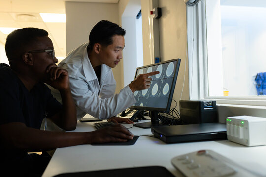 Diverse male medical staff wearing lab coat and scrubs reviewing brain scans on workstation monitor