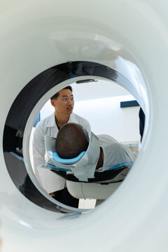 Asian tech in lab coat adjusting patient in hospital gown inside scanner gantry on blue cushion