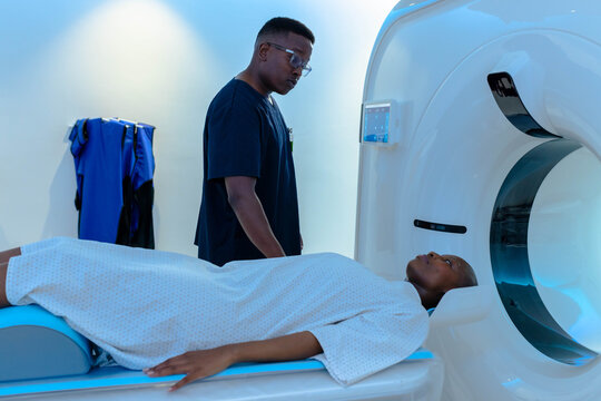 African American tech in scrubs with patient in gown preparing for CT scan near gantry, aprons
