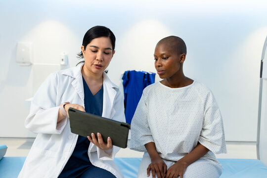 Diverse female provider in white lab coat discussing tablet with patient sitting on exam table