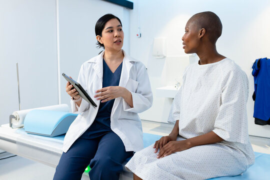 Female clinician in lab coat, female patient in gown sitting on exam table discussing with tablet