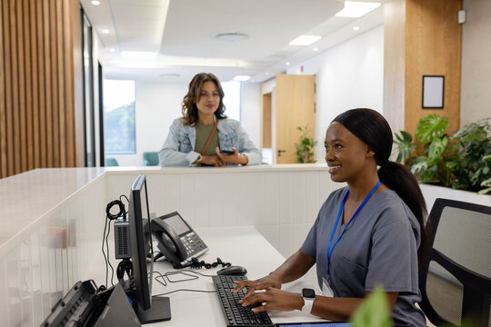 Female receptionist in scrubs typing on computer at desk, visitor holding phone, strap, copy space