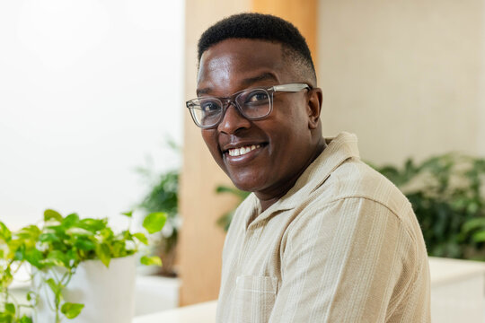 Adult African American man smiling in lounge in tan shirt and clear glasses beside white planter