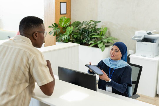 African American hijab receptionist showing tablet and lanyard to male visitor at desk, copy space