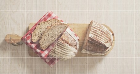 Showing two rustic sourdough loaves resting on light wood table, with cutting boards, gingham cloth