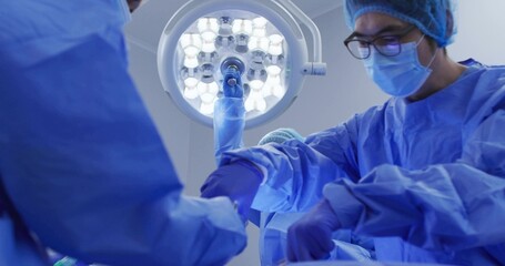 Operating surgical team wearing blue gowns, masks, gloves, working in operating room with LED light