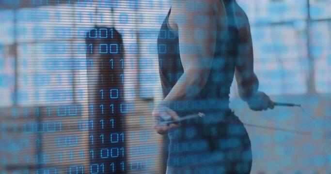 Jumping woman wearing dark workout gear holding jump-rope in gym, with heavy bag and binary overlay - Powered by Adobe