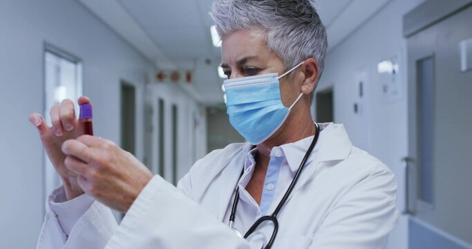 Holding doctor inspecting purple capped tube in clinic corridor, white coat blue mask stethoscope