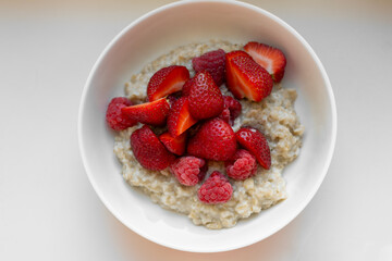 Top-down view of creamy oatmeal served in a white ceramic bowl, topped with halved ripe strawberries and whole raspberries on a white table. The vibrant red berries with the soft beige oats.