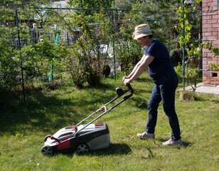 An older woman in straw hat, working in the spring, mows the grass in her backyard. Concepts of gardening, work, and nature. Concept of movement in adulthood. Lawn maintenance before the season.