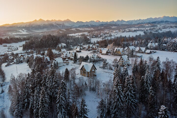 Atmospheric aerial view of a snowy village in Białka Tatrzańska, Poland, nestled among frosted forests just before sunrise in December, with the Tatra Mountains in the distant background.