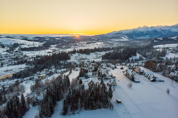 Golden hour sunrise view over the snowy village of Białka Tatrzańska in December, with the warm light illuminating the forest, traditional architecture, and the majestic Tatra Mountains.