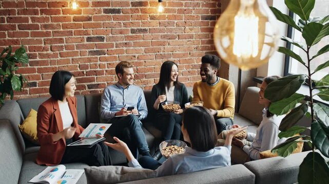 Diverse group of young business colleagues relaxing on a modern couch during a break, eating snack food and reviewing documents in a loft office