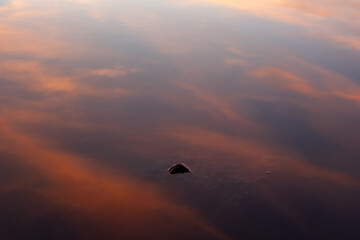 A single rock breaks the lake’s surface, mirroring the sunset sky in soft hues of orange and blue.