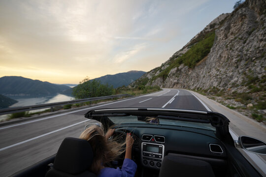Young woman driving convertible along scenic ocean coast and mountain road at sunset, carefree summer vacation, travel freedom and adventure on coastal highway - Powered by Adobe