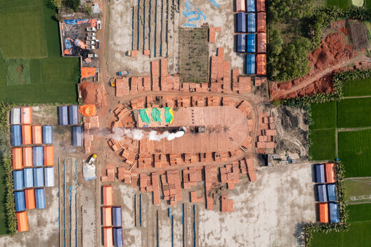 Aerial view of an oval brick kiln with smoke rising amidst a grid of green fields and stacks of colorful bricks, Bogura, Rajshahi Division, Bangladesh.