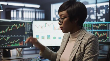 Focused Black woman working with stock charts and financial data on computer screen, analyzing business trends in modern office. - Powered by Adobe