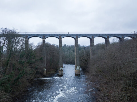 Aerial view of a majestic stone viaduct piercing the sky above a rushing river, framed by the stark beauty of winter trees, Llangollen, Wales, United Kingdom.