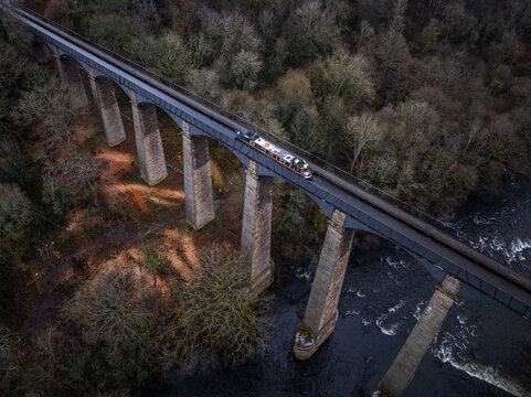 Aerial view of a vintage train traversing the majestic Pontcysyllte Aqueduct, a ribbon of steel and stone amidst a tapestry of dark forest and the river below, Llangollen, Wales, United Kingdom.