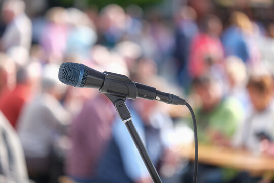 Seitliche Nahaufnahme eines Mikrofons im Halter vor einer Masse an unkenntlichen Menschen im Publikum im Hintergrund auf einer Open-Air Veranstaltung &ndash; selektiver Fokus, sehr viel Tiefenunsch&auml;rfe