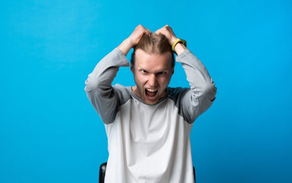 Angry young man pulling his hair in frustration on blue background, emotional stress and rage expression