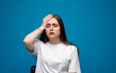 Stressed young woman holding head with hand, tired emotional facial expression on blue studio background