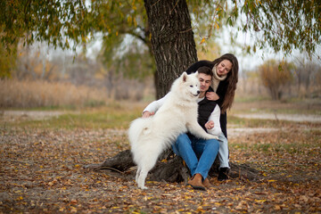 A young family - a guy, a girl and a cute fluffy Samoyed - are walking by the river