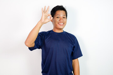 A cheerful young Asian man in a blue shirt stands against a clean white background, smiling and giving an OK gesture.