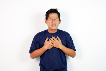 A young Asian man in a blue shirt clutches his chest with a pained expression, conveying distress or chest discomfort in a simple studio setting.