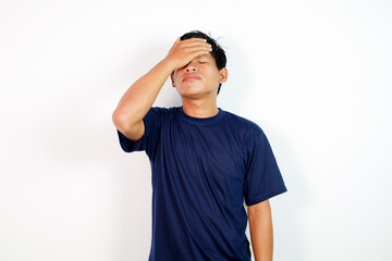A young Asian man wearing a blue shirt rests his hand on his forehead, signaling stress or fatigue. He stands against a clean white backdrop, conveying worry, confusion, or exhaustion.