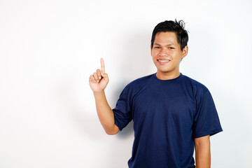 A Cheerful Young Asian Man Wearing a Blue Shirt Pointing Upward With a Smile on a White Background.