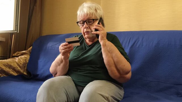 Senior woman sitting on sofa holding credit card and talking on phone. Concept of banking fraud protection, phone scam, customer service, remote payment