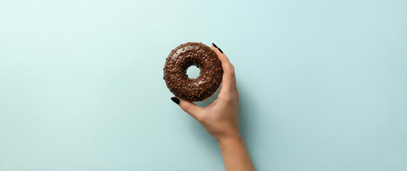 Female hand with black nail polish holding a delicious chocolate glazed donut with sprinkles. Minimalist food photography concept on a pastel blue background with copy space