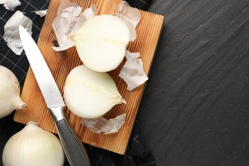 Fresh onion bulbs with peels and knife on black textured table, flat lay. Space for text