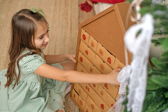 A cute beautiful girl in a green dress opens a cell of the Advent calendar, sitting by the Christmas tree. Looking forward to the New Year and Christmas