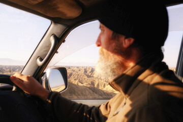A man with a gray beard is driving a car. A dry mountain landscape is visible from the window. Sunlight falls on the driver's face