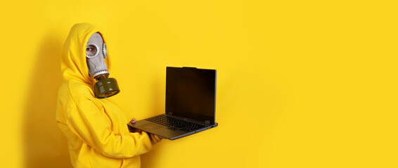 Individual in yellow hoodie and gas mask holds open laptop against vibrant yellow background. Modern, conceptual image with copy space.