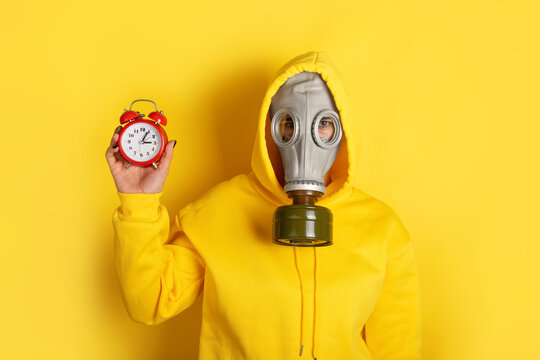 Individual wearing yellow hoodie and gas mask holds red alarm clock, standing against vibrant yellow background. Concept of urgency, danger, and protection.