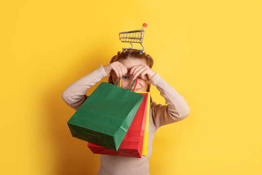 Cheerful girl with colorful shopping bags and a toy cart on her head against a vibrant yellow background, symbolizing retail fun and creativity.